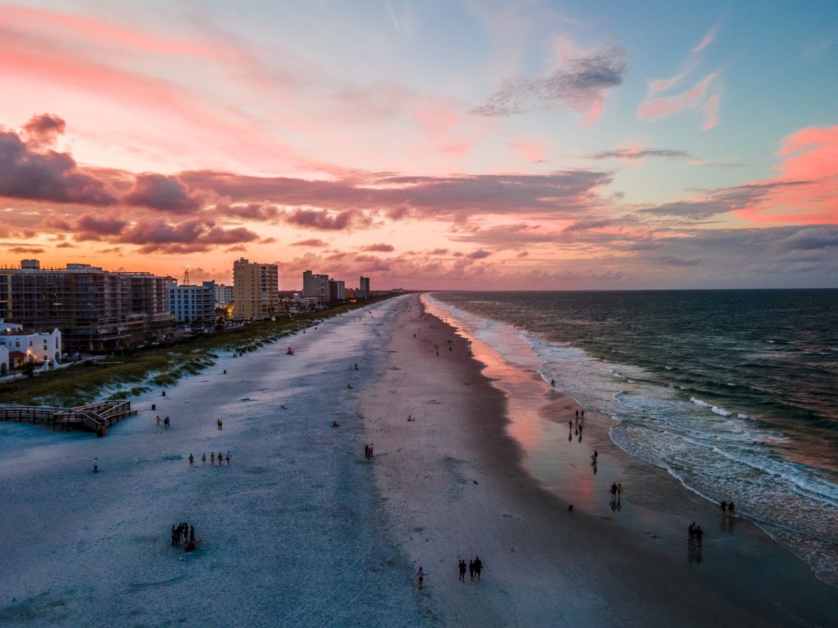 A beautiful beach at sunset with a colorful sky, ocean waves, and people walking along the shore. Buildings line the coast in the distance.