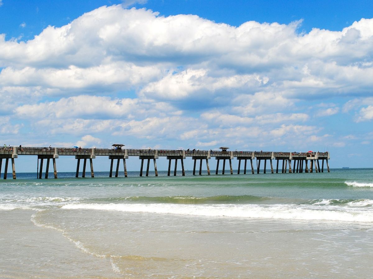 A long pier extends over the ocean against a backdrop of fluffy clouds and a sandy beach.