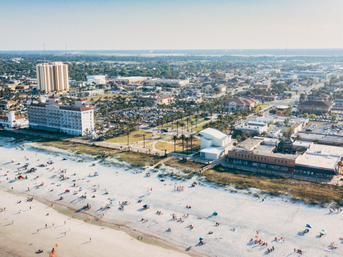 Aerial view of a sandy beach with people, adjacent to a coastal city with buildings and greenery under a clear sky.
