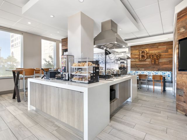 The image shows a modern kitchen or cafe area with tables, a counter with food items, and stylish decor on wooden walls.