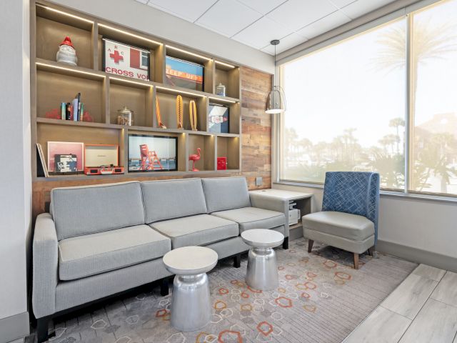The image shows a modern waiting room with a gray sofa, blue chair, small tables, and a shelf with decorative items, near a large window.