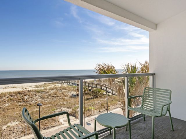 A coastal balcony view with chairs and a table, overlooking a beach and ocean under a clear blue sky.