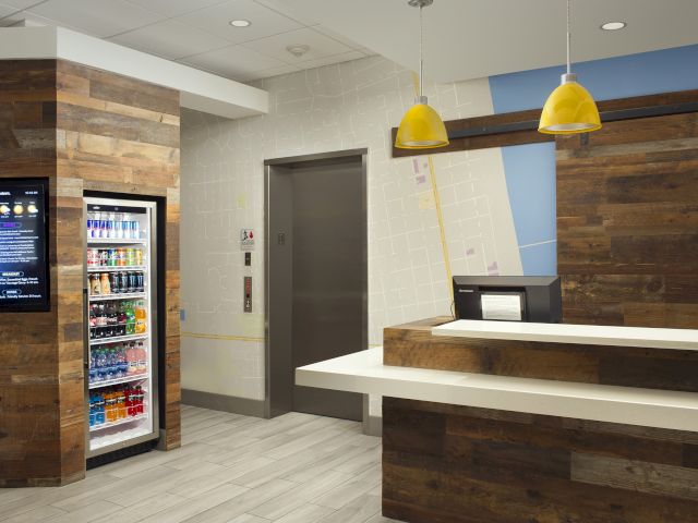The image shows a modern hotel lobby with wooden accents, a snack bar, a drink fridge, pendant lights, and an elevator.