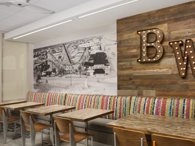 The image shows a cozy cafe with colorful seating, wooden tables, and a wall with a lit sign and a black-and-white mural.