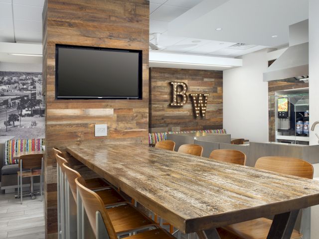 The image shows a stylish dining area with a wooden table, chairs, a TV on the wall, and decorative signage, all in a modern design.