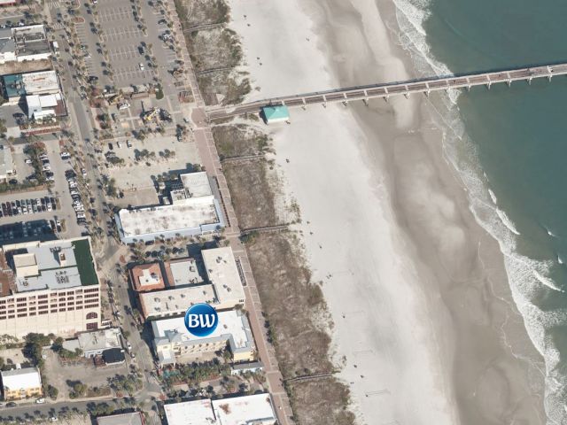 An aerial view of a coastal area shows a beach, a pier extending into the water, and urban buildings nearby, including one with a circular logo.