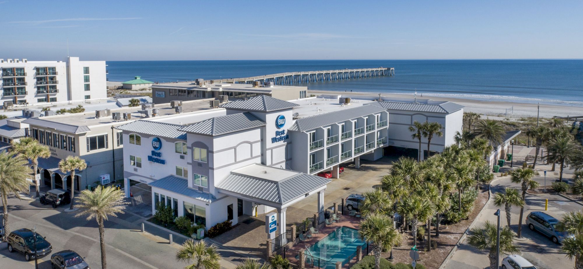 A coastal area features hotels, a pool, palm trees, and a long pier extending into the ocean under a clear blue sky.