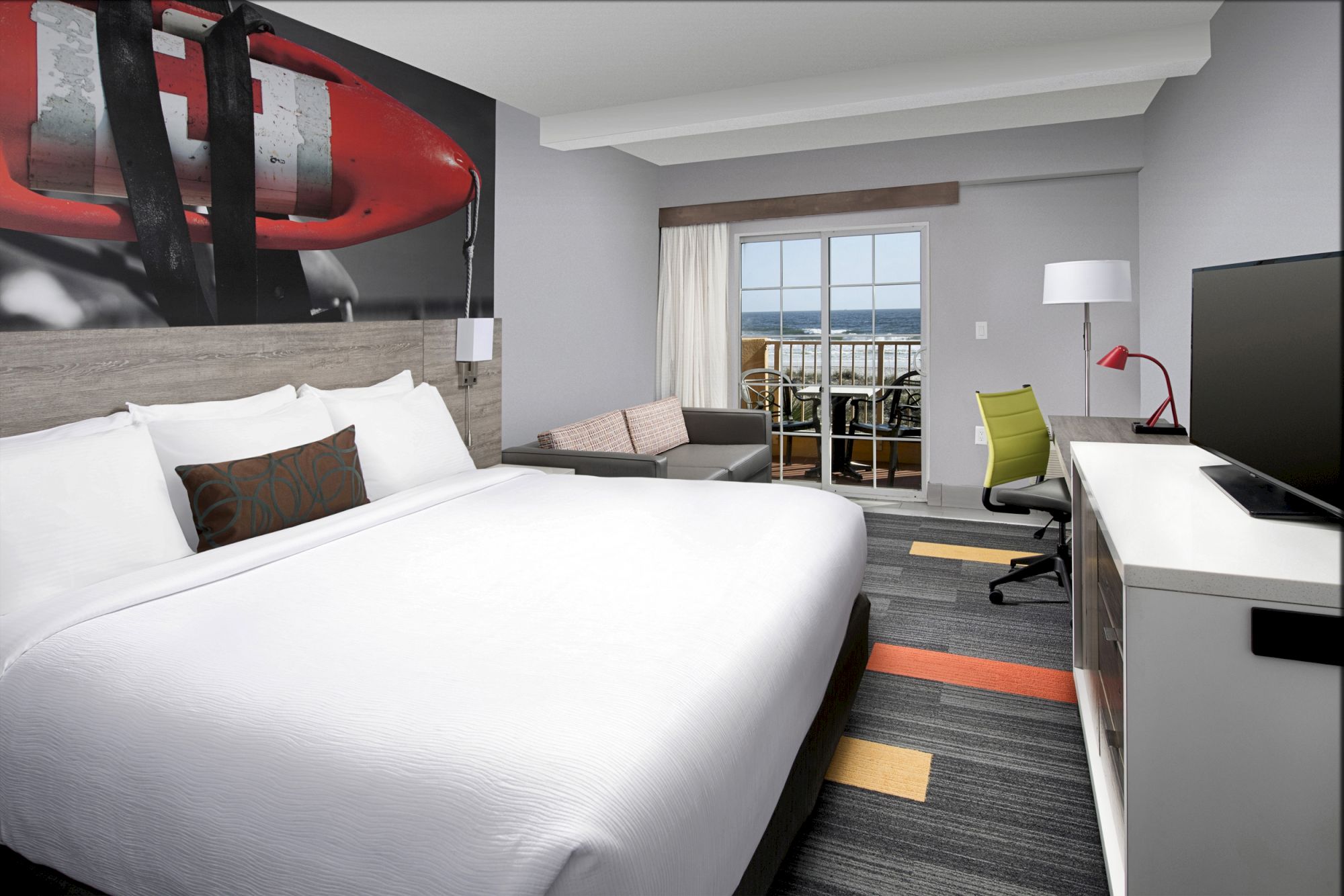 The image shows a modern hotel room with a large bed, TV, desk, chair, and view of a balcony through sliding glass doors.