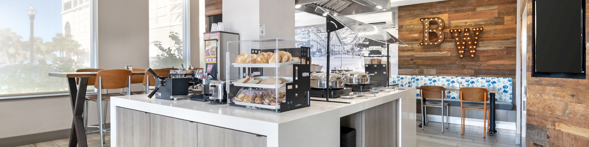 The image shows a modern cafe with a central counter, displaying baked goods and seating. Wood accents and light colors create an inviting space.